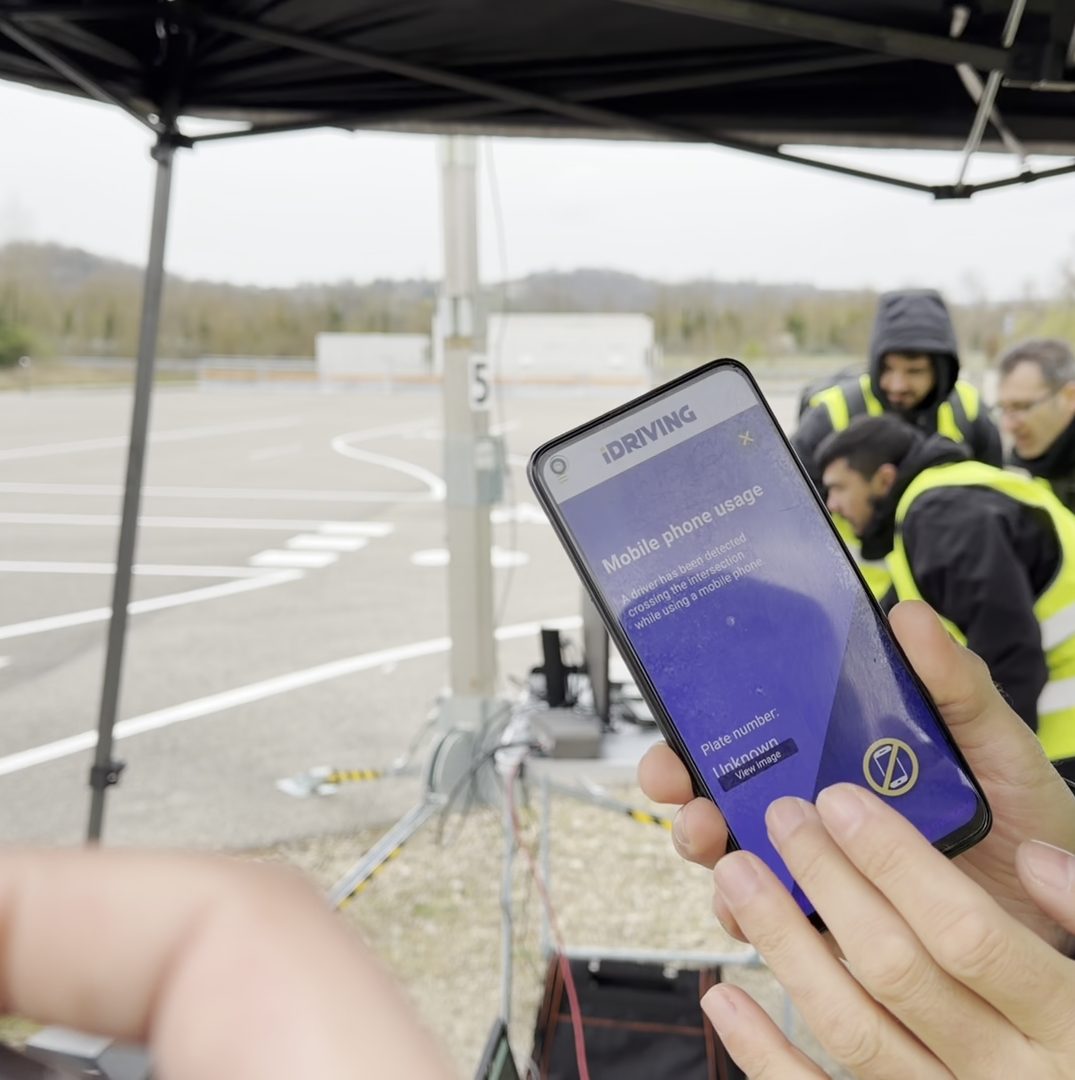 A smartphone shows an iDriving app alert about mobile phone use detected at the test intersection